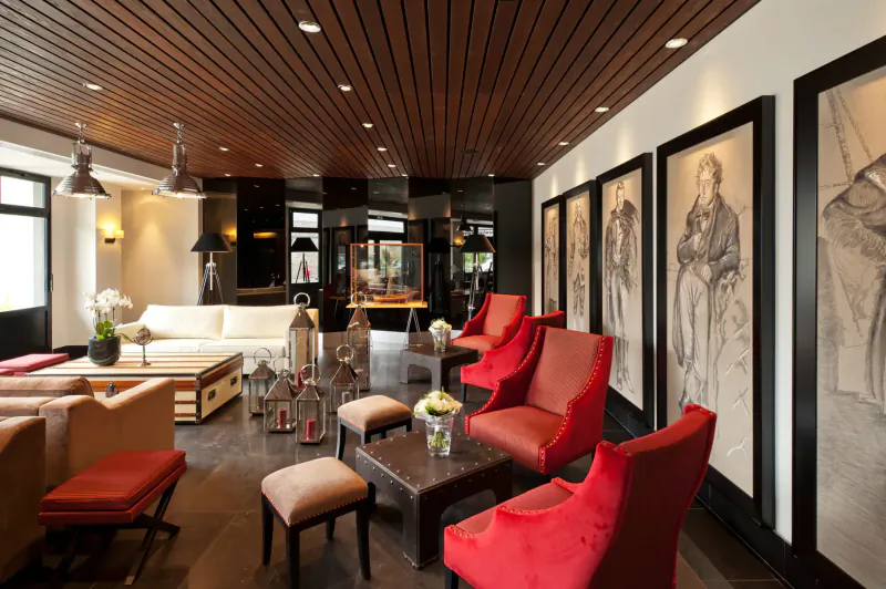 Elegant Nouveau Monde gallery lounge in Saint-Malo with wooden ceiling, red armchairs, white sofas, lanterns, and large black-framed art.