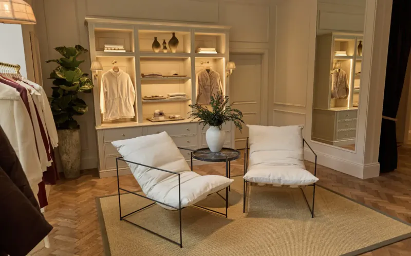 Elegant Varley store interior in SoHo: white lounge chairs, glass table with flowers, lit display cabinets with folded clothes.