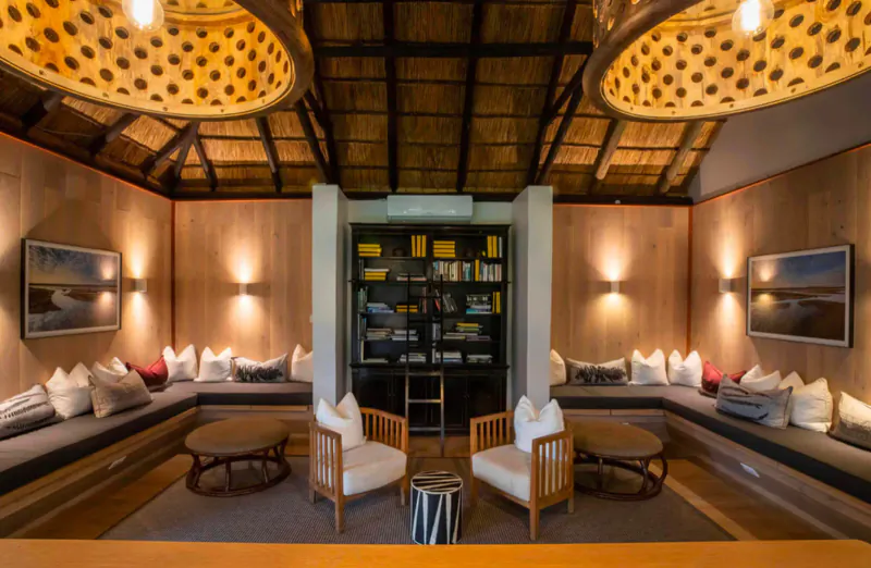 Cozy lounge in Mfuwe Lodge with thatched ceiling, woven pendant lights, bookshelves, white chairs, and plush cushions.