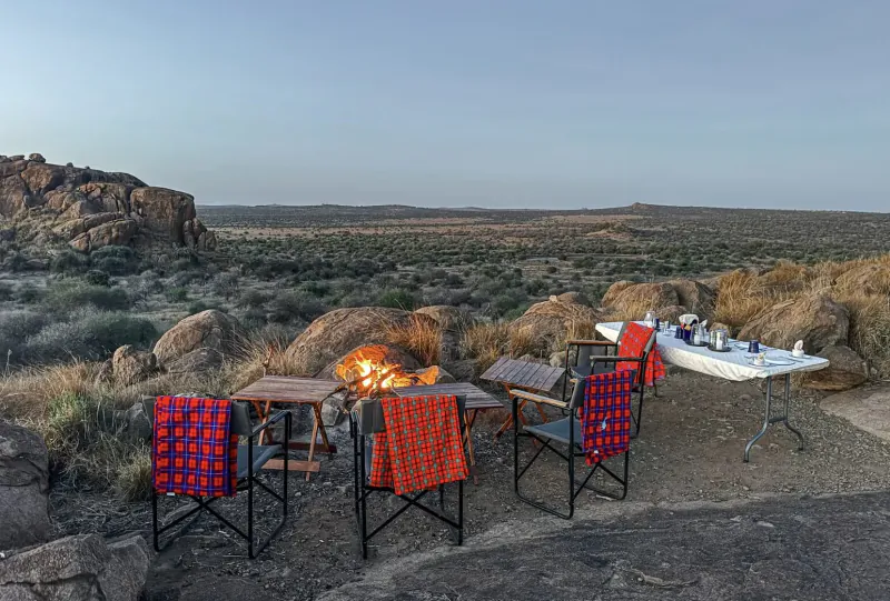Campfire with orange blankets on chairs and folding table with drinks at Tumaren Camp, rocky savanna at dusk.