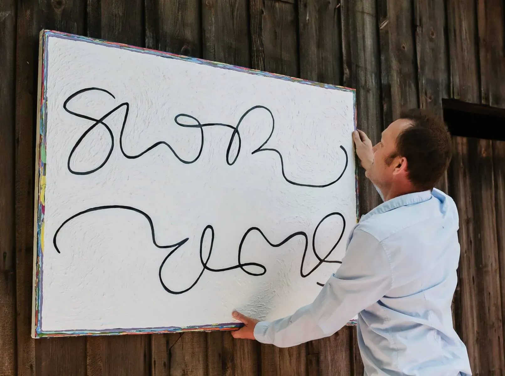 Man in light blue shirt hanging white-framed sign reading 'Snowmore' on wooden wall
