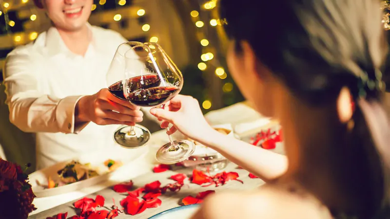 Smiling man in white shirt toasts red wine with woman at candlelit table with rose petals, string lights backdrop.