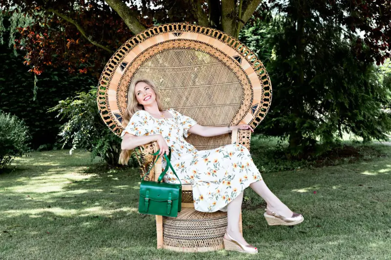 Blonde woman lounging in large woven rattan chair on lawn, wearing floral dress and holding green satchel bag.