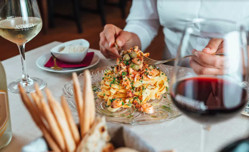 Close-up of woman eating shrimp pasta topped with prawns at Italian restaurant table with white wine, red wine, and grissini.