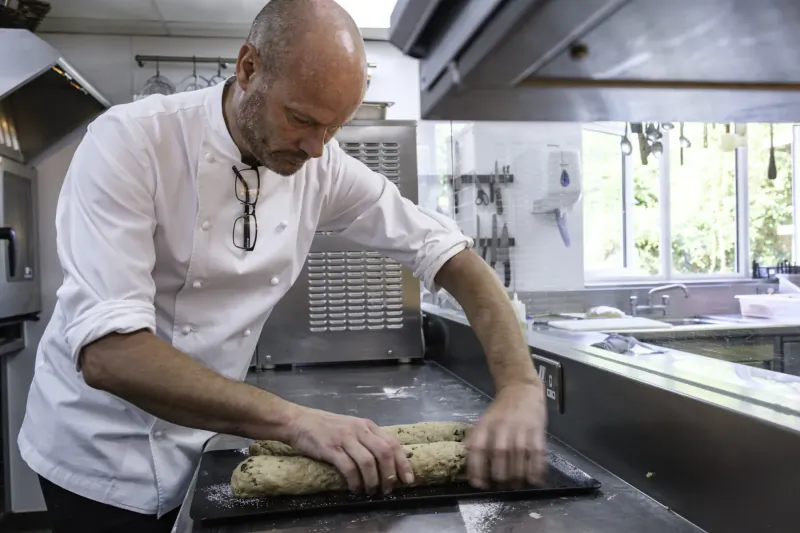 Bald chef in white uniform shaping log-shaped dough on board in professional kitchen