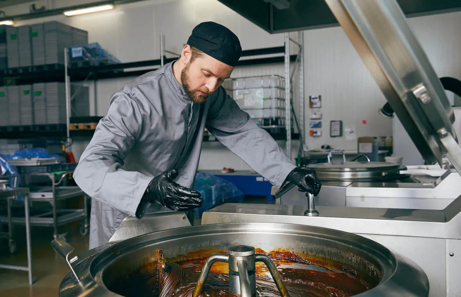 Bearded chef in gray uniform and black cap stirring thick red sauce in large industrial mixer in kitchen
