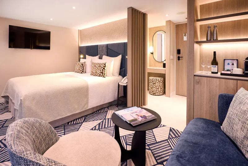 Spacious VIVA ENJOY cruise ship suite with king bed, blue sofa, coffee table, TV, and minibar.