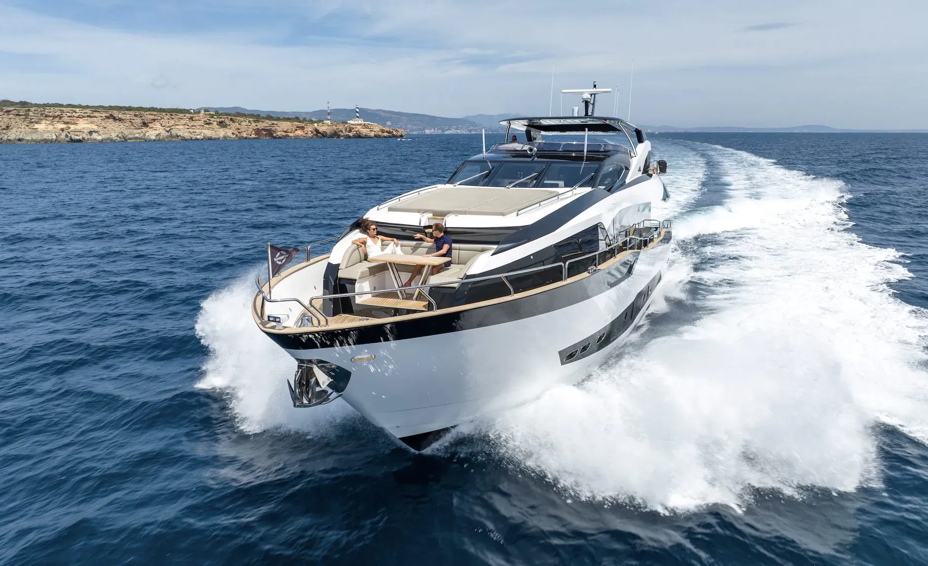 White luxury yacht speeding on blue sea with white wake, people seated on deck, rocky coastal backdrop.