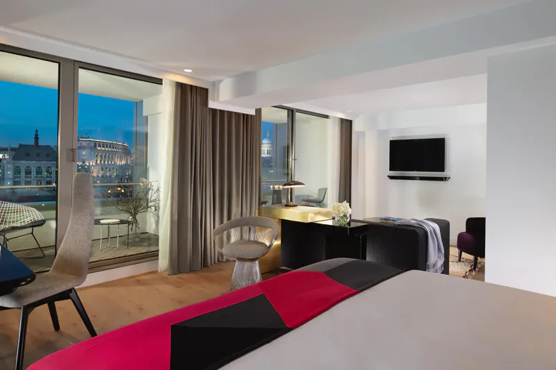 Modern luxury hotel room at dusk with panoramic city view, plush bed, desk, chairs, and TV.