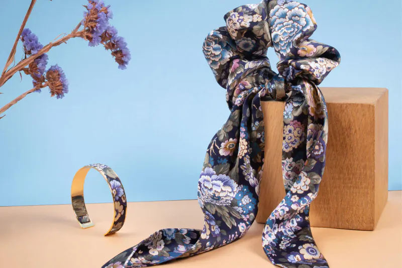Floral navy scrunchie draped on wooden block with matching gold bracelet, against blue backdrop with lavender stems