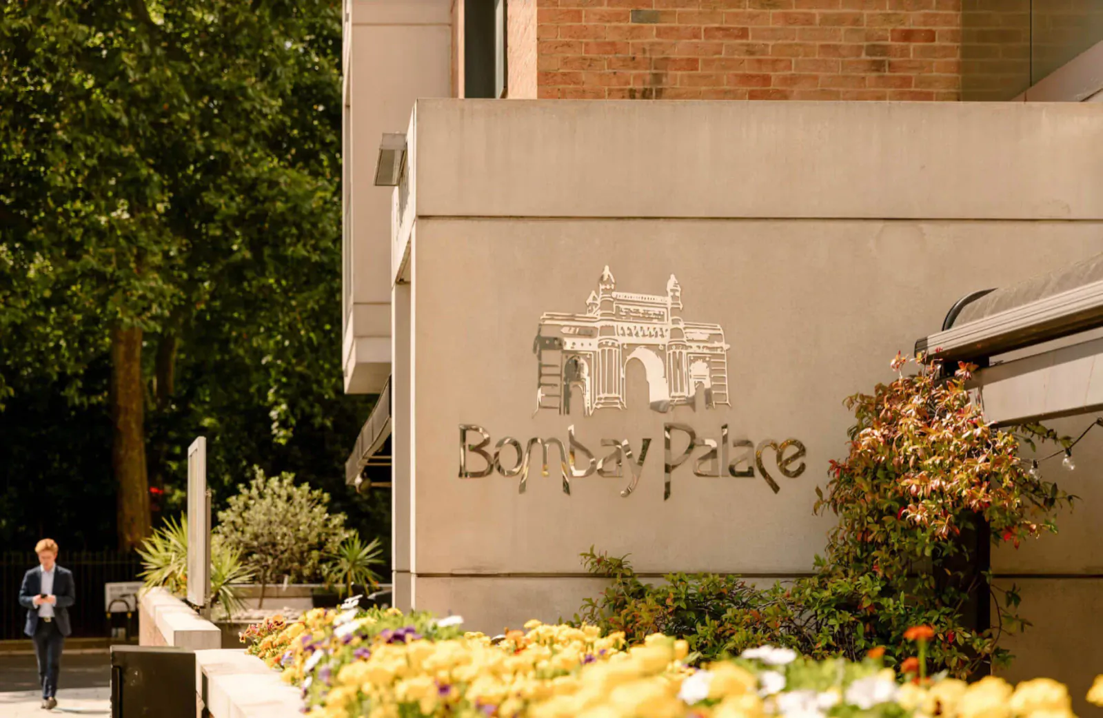 Bombay Palace building facade with arched logo, yellow flowers, greenery, and man in suit walking nearby