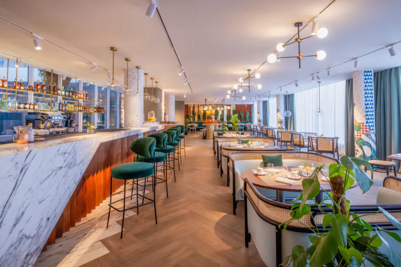 Modern hotel bar and dining area at Gem Forest Hotel, Nairobi, with marble bar, green velvet chairs, plants, and pendant lights.