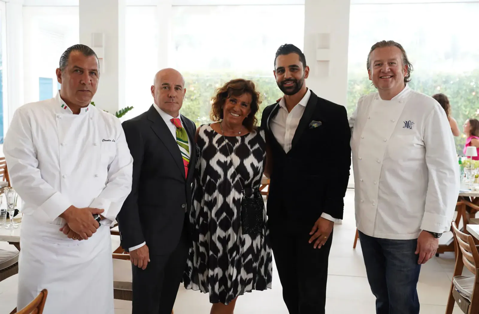 Four people posing together at Italian affair event: two chefs in white uniforms, man in suit, woman in patterned dress, sunny dining area.