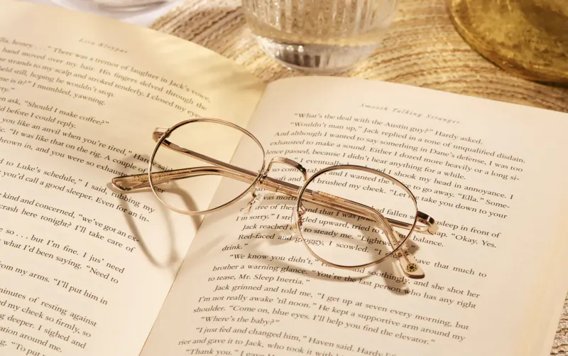 Open books on table with gold wire-rimmed glasses resting across them, water glass and gold vase nearby.