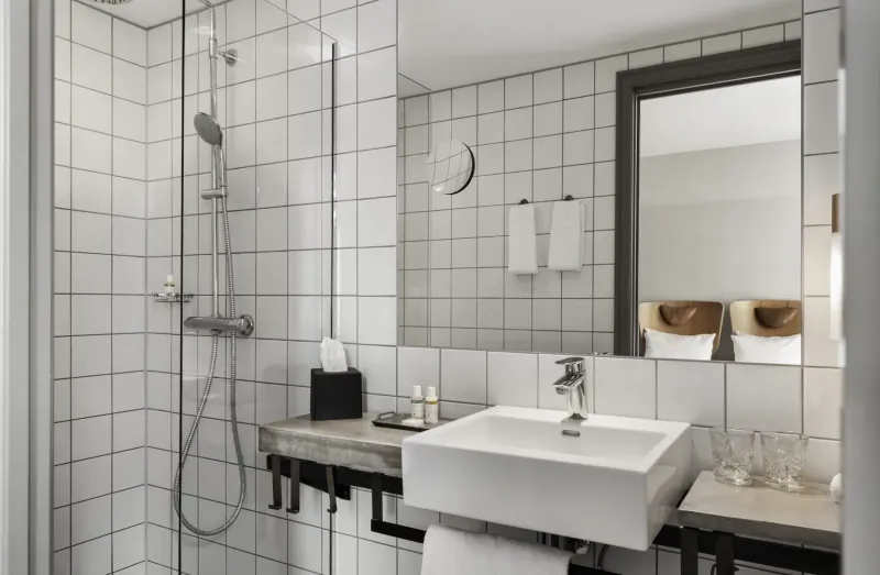 Modern white-tiled hotel bathroom with glass shower, sink, mirror, towels, and amenities in Copenhagen's Hotel SP34.