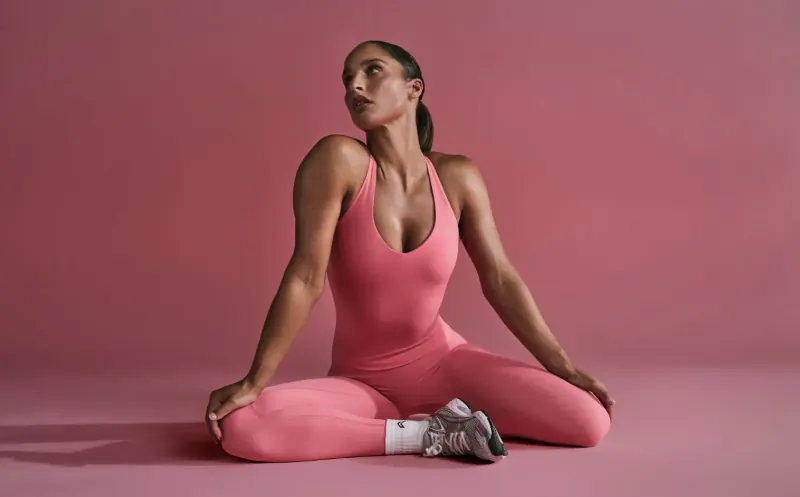Athletic woman in pink Oner Active bodysuit sits cross-legged, looking up, on pink background.