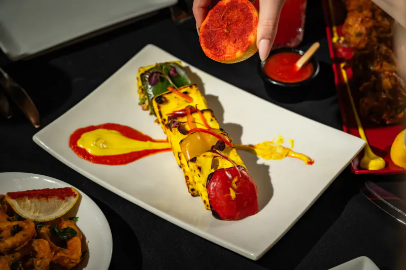 Grilled vegetable skewer on white plate with red and yellow sauces, hand holding blood orange slice, at Mint Leaf London