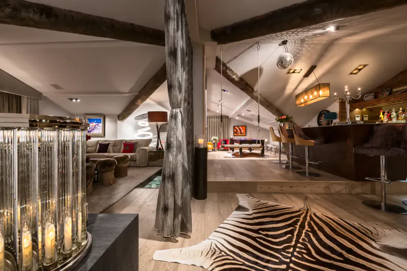 Luxurious mountainside chalet interior with wooden beams, zebra rug, pool table, bar, and modern lounge.