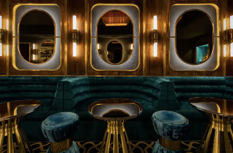 Elegant Disrepute Bar interior in Soho: teal velvet curved booth, gold pedestal tables and stools, oval lit mirrors.