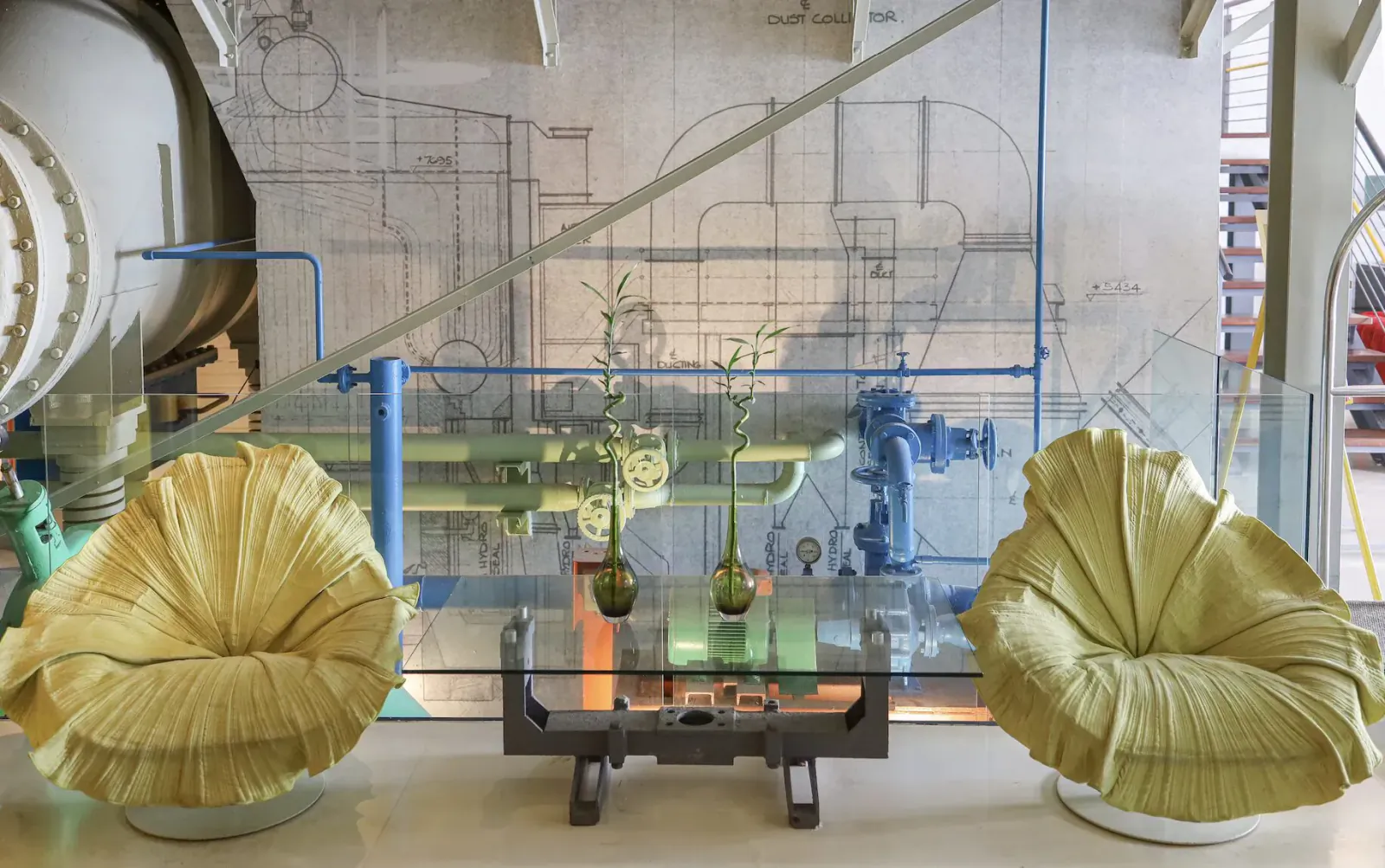 Two yellow leaf-shaped armchairs with plants flank a metal coffee table in a luxury hotel's industrial turbine room.