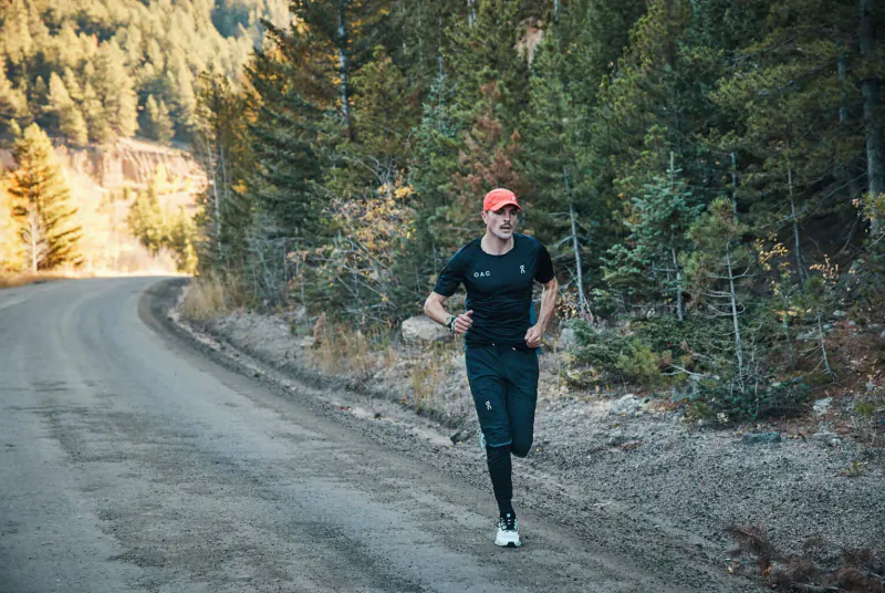 Runner in orange cap and black outfit jogging on rural dirt road through pine forest, 'Run for each other' event.