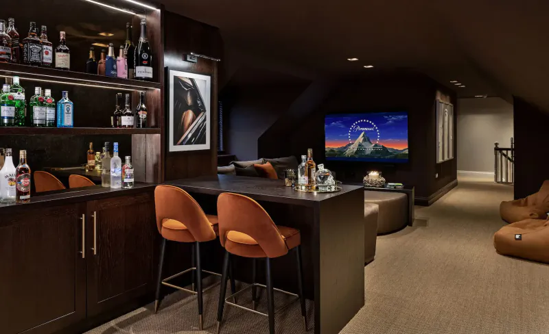 Elegant attic bar with backlit wood shelves stocked with liquor bottles, orange swivel stools, TV on mountain landscape, and lounge seating.