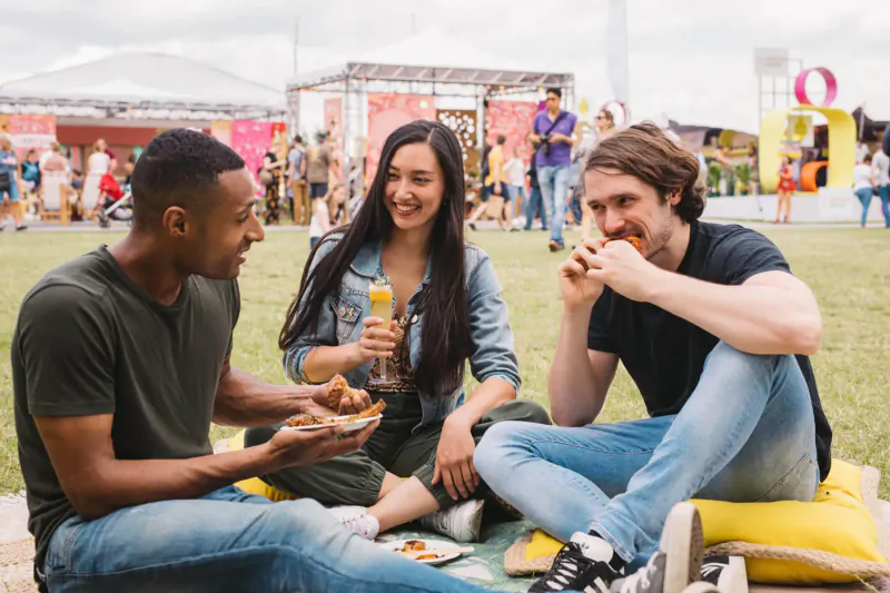 Three diverse friends eating pizza and snacks, laughing on a picnic blanket at Taste of London festival amid crowds.