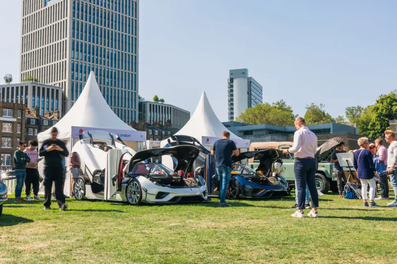 Outdoor Concours d'Elegance car show with luxury vehicles like white supercars under tents, crowds viewing, skyscrapers backdrop.