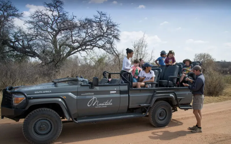 Group of tourists in gray Marataba safari vehicle on dirt road amid African bushveld savanna under cloudy sky