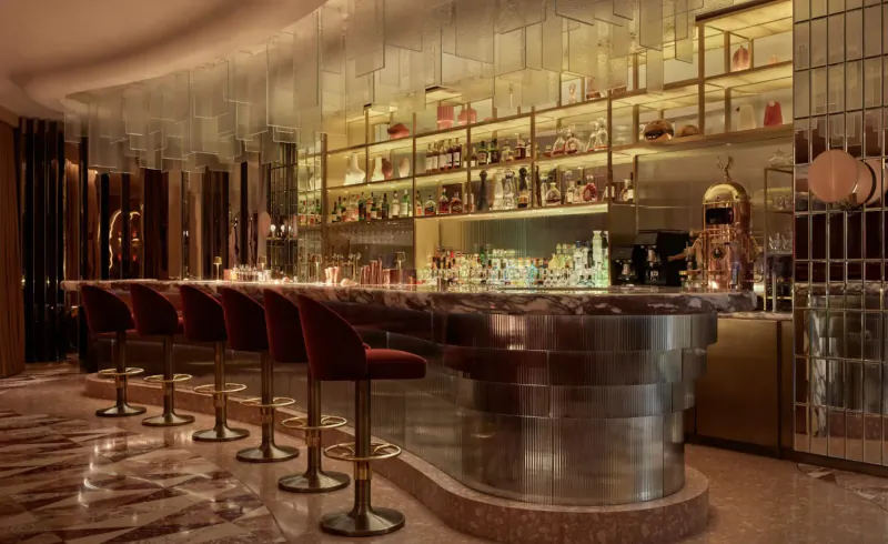 Elegant Revery Bar with curved metallic bar, red velvet stools, backlit bottle shelves, and afternoon tea setup in luxurious setting