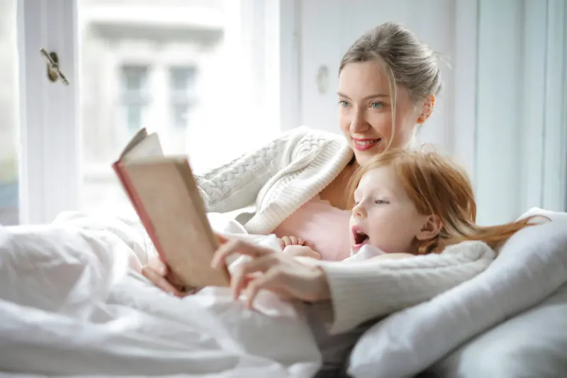 Mother in white sweater reading book to red-haired girl in pink pajamas in sunlit bed