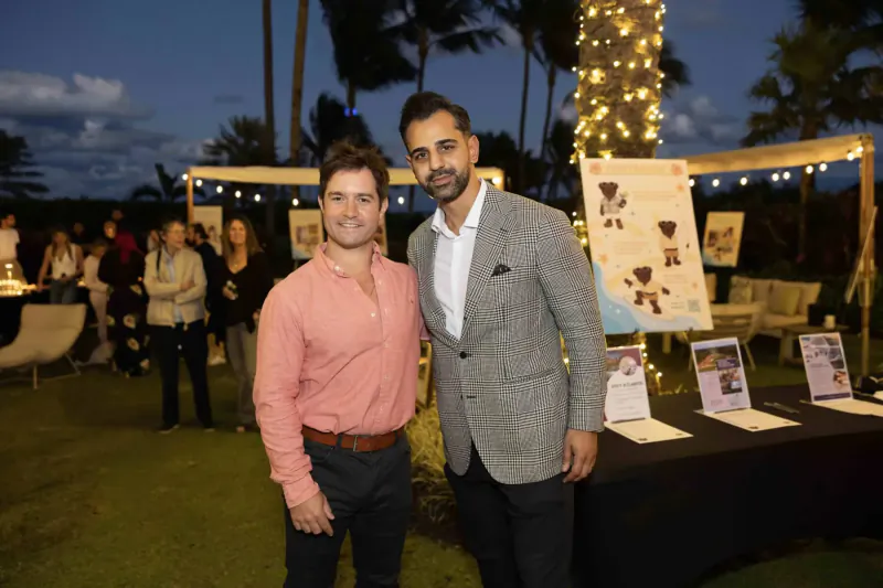 Two men in suits pose smiling at outdoor evening charity event for Childhood Cancer Society with palm trees and display boards.