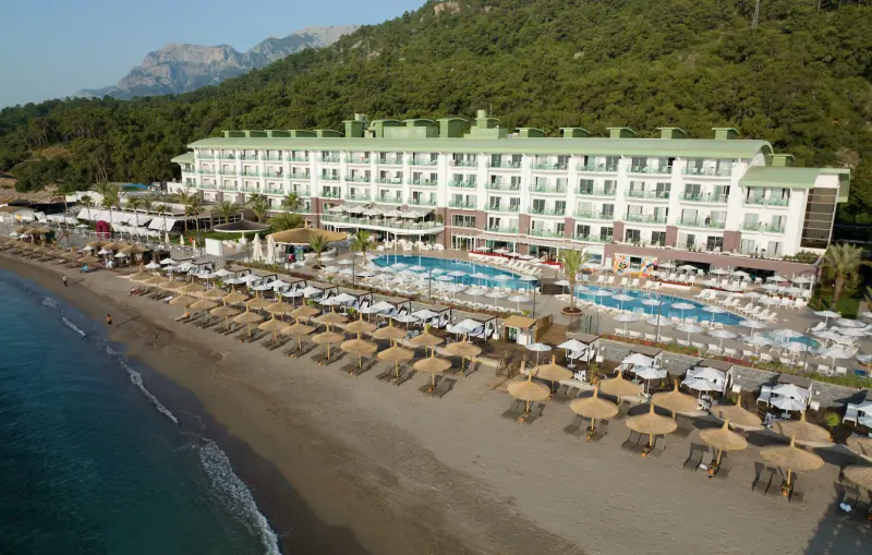 Aerial view of Corendon Playa Kemer luxury resort with pools, beach umbrellas, and sea against green mountains.