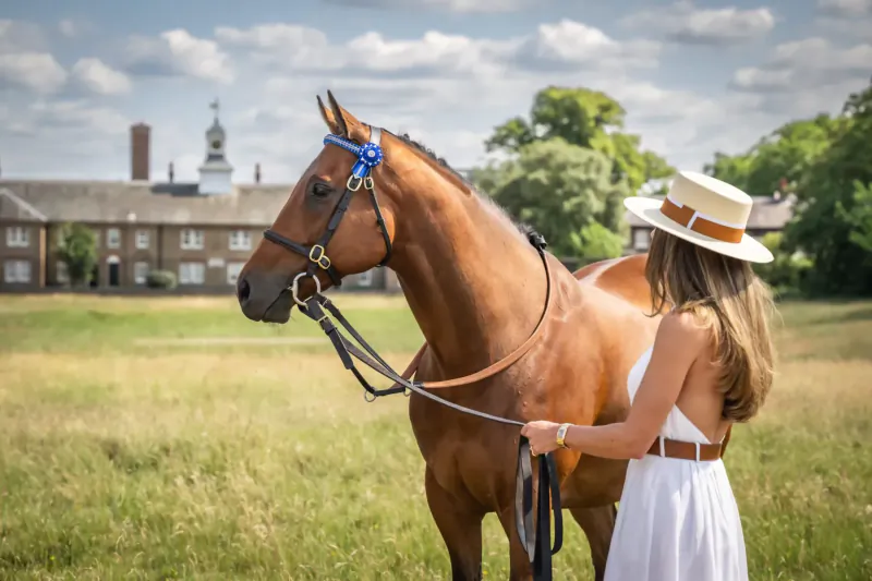 Woman in white dress and straw hat holds reins of bay horse with blue bridle in grassy field before historic buildings.