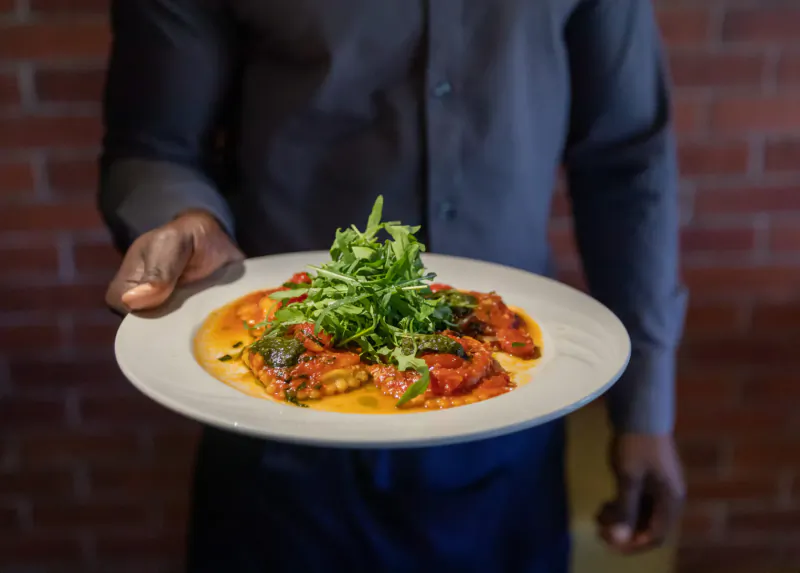 Server holding white plate of tomato sauce pasta topped with arugula against brick wall
