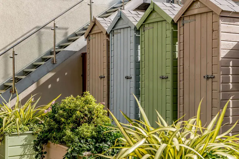 Row of colorful wooden beach huts in shades of green, blue, tan beside metal stairs and potted plants at Woolacombe Bay