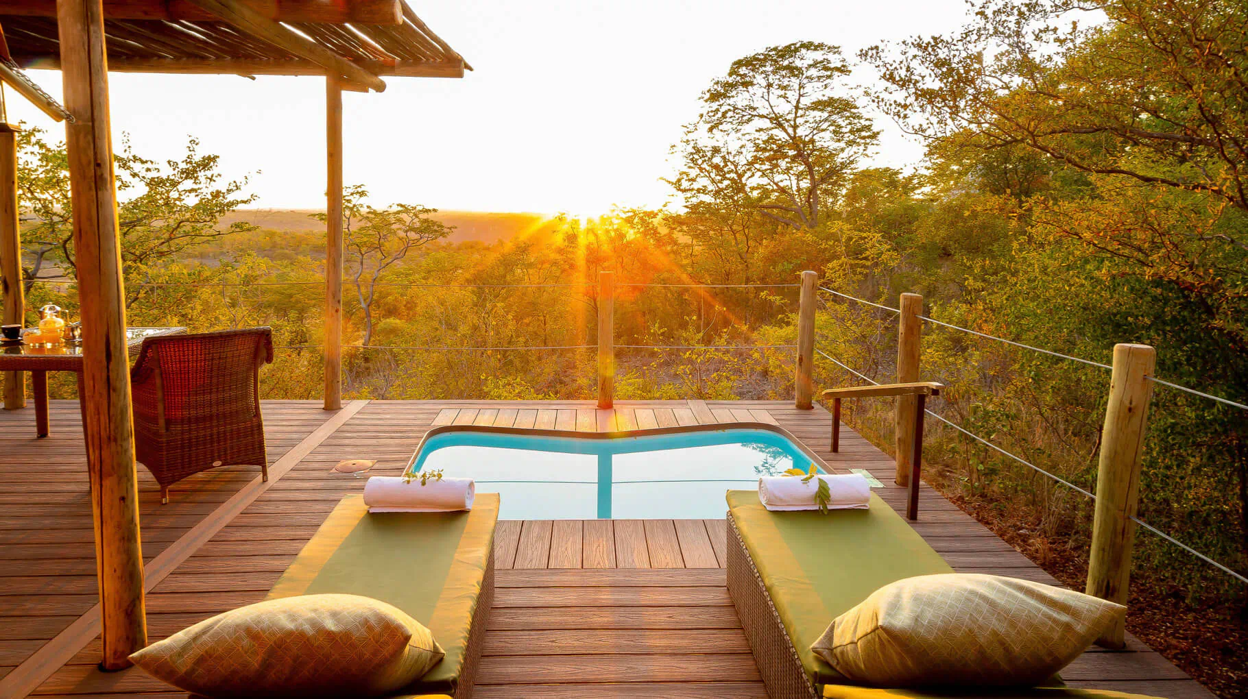 Sunset view from wooden deck at Old Drift Lodge: infinity pool with towels, loungers, bushveld landscape.