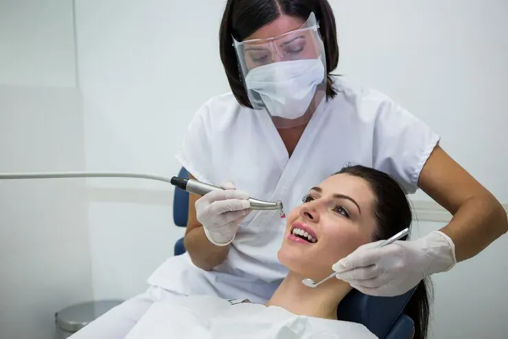 Dentist in PPE using drill on smiling female patient in dental chair during cosmetic treatment
