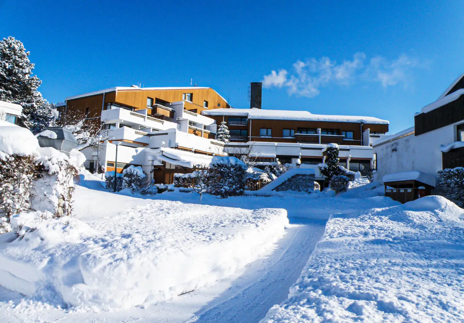 Snowy winter scene at Karma Bavaria resort in Schliersee, Germany, with snow-covered buildings and pine trees under blue sky.