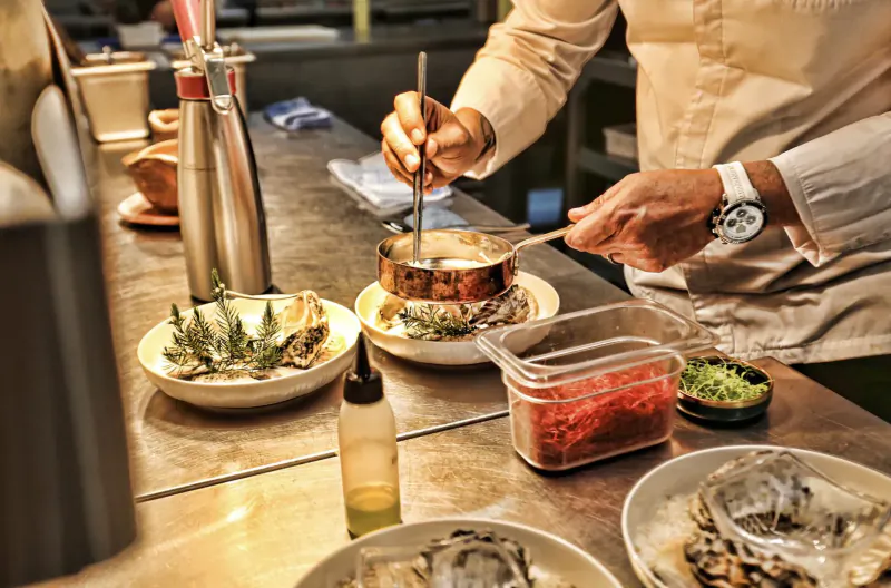 Chef in beige uniform stirring copper pot of oysters and garnishes on professional kitchen counter