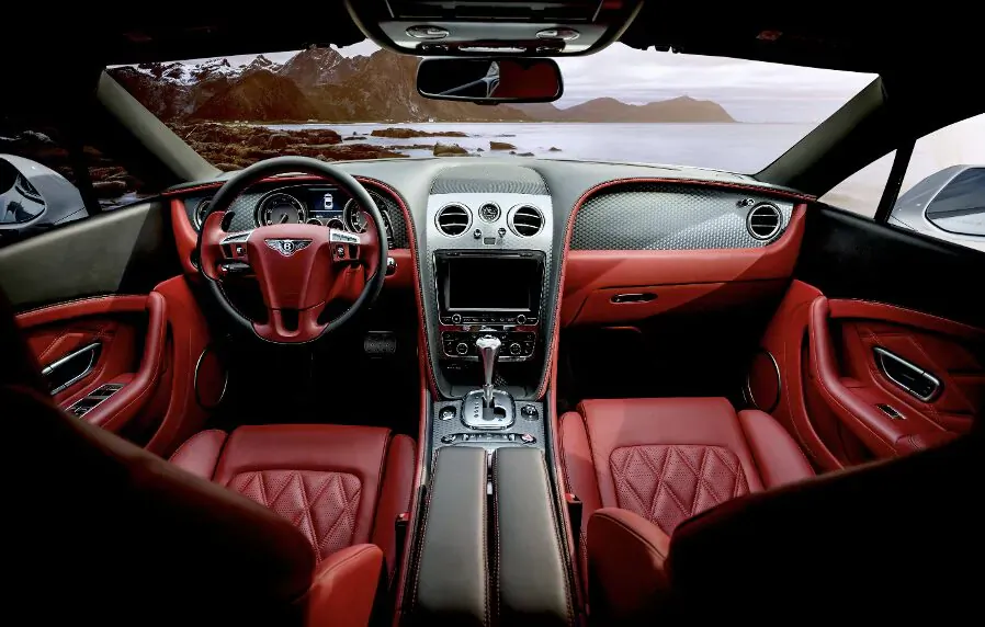 Luxury Bentley Continental GT interior with red leather seats, steering wheel, dashboard, viewed from driver's seat facing scenic lake.