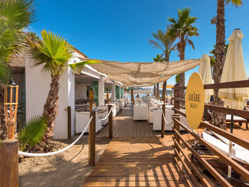 Amàre Beach entrance at Marbella beach club: shaded wooden deck with white loungers, palms, umbrellas under blue sky.