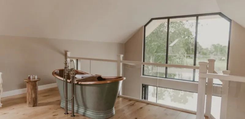Freestanding copper bathtub on wooden floor in loft bathroom with large window overlooking lake at The Boathouse.