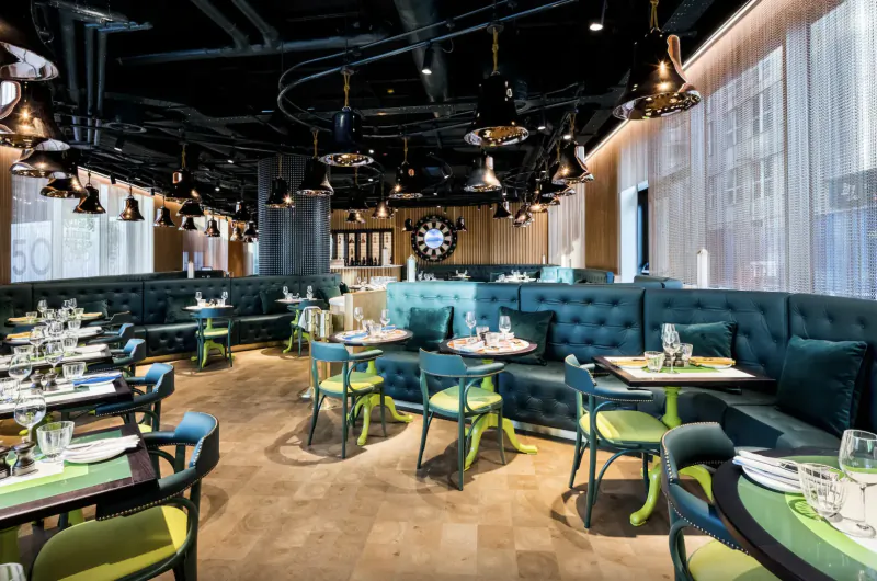 Modern hotel restaurant interior with teal booths, green chairs, wooden floors, pendant lights, and large clock.