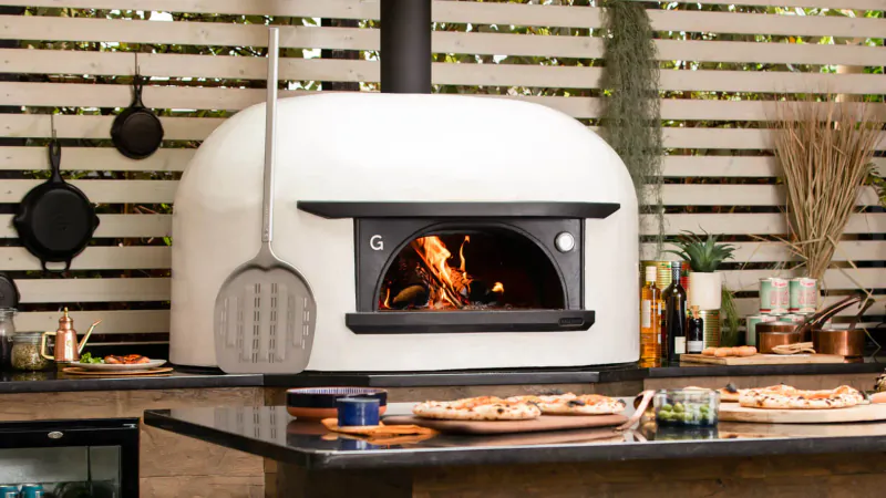 White Gozney wood-fired pizza oven with flames, peel, pizzas on outdoor wooden counter amid garden decor.
