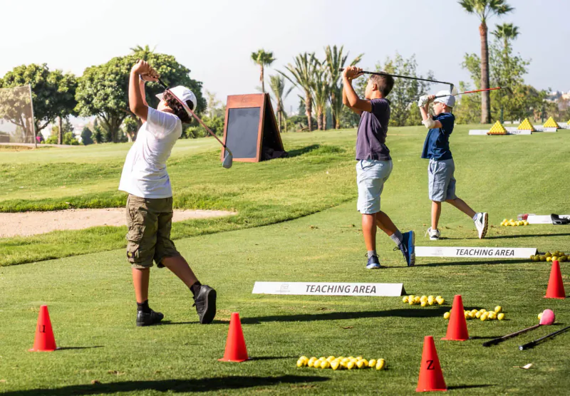Three boys practicing golf swings in Los Naranjos teaching area with cones, balls, and whiteboard on green course.