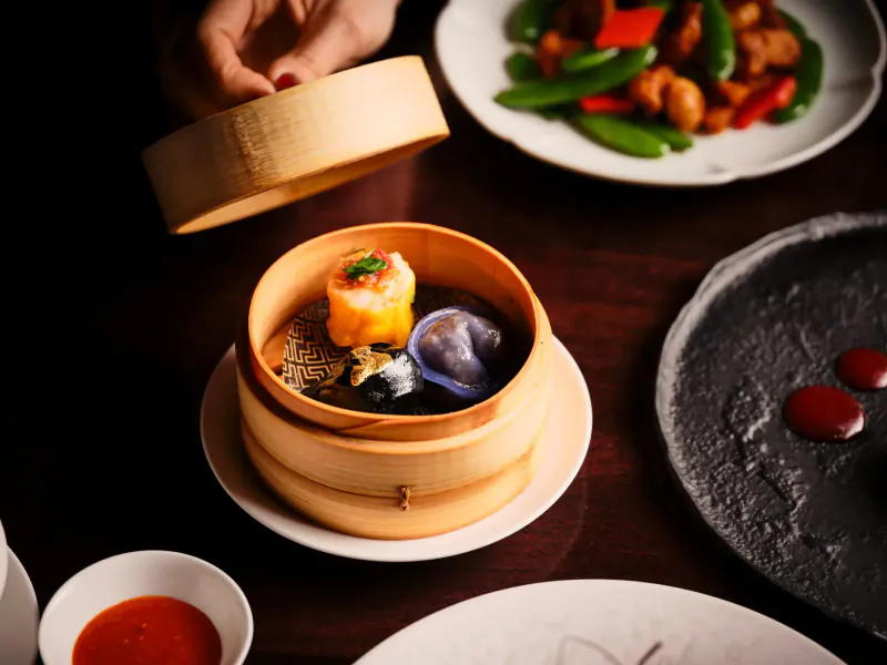 Hand lifting lid from bamboo steamer revealing orange, purple dumplings; side dishes of shrimp and peppers, chili sauce on wooden table