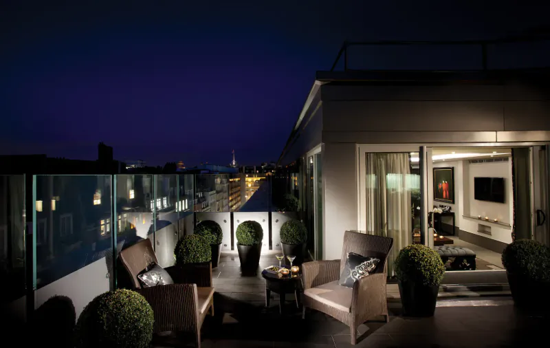 Luxurious rooftop terrace at night with lounge chairs, plants, drinks, and city skyline view at The May Fair Hotel.