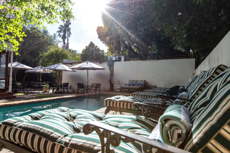 Sunny poolside at Evergreen Manor and Spa, Stellenbosch, with striped lounge chairs, umbrellas, and lush trees.