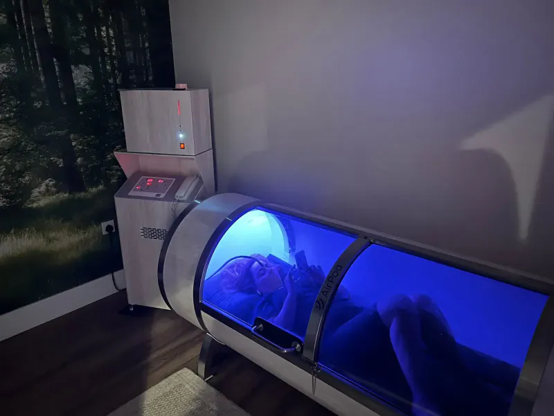 Person reclining in blue-lit cylindrical Aqua-Tox pod at wellness clinic with forest wall mural